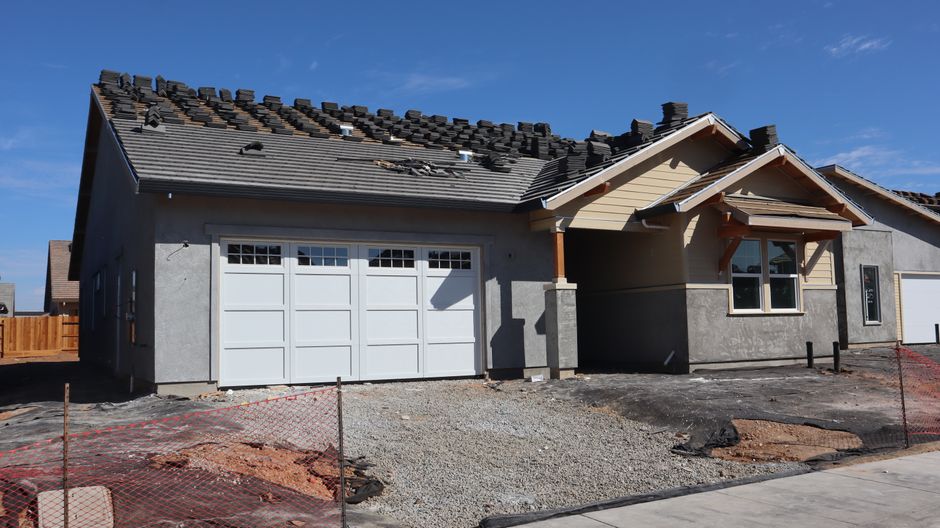 New single-story house under construction with a white garage door, gray stucco walls and roof tiles stacked on the roof